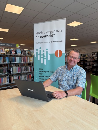 Vrijwilliger Dik zit aan tafel klaar om vragen te beantwoorden. Voor hem staat een laptop om zaken op te zoeken. Achter hem staat een roll-up banner van het IDO.